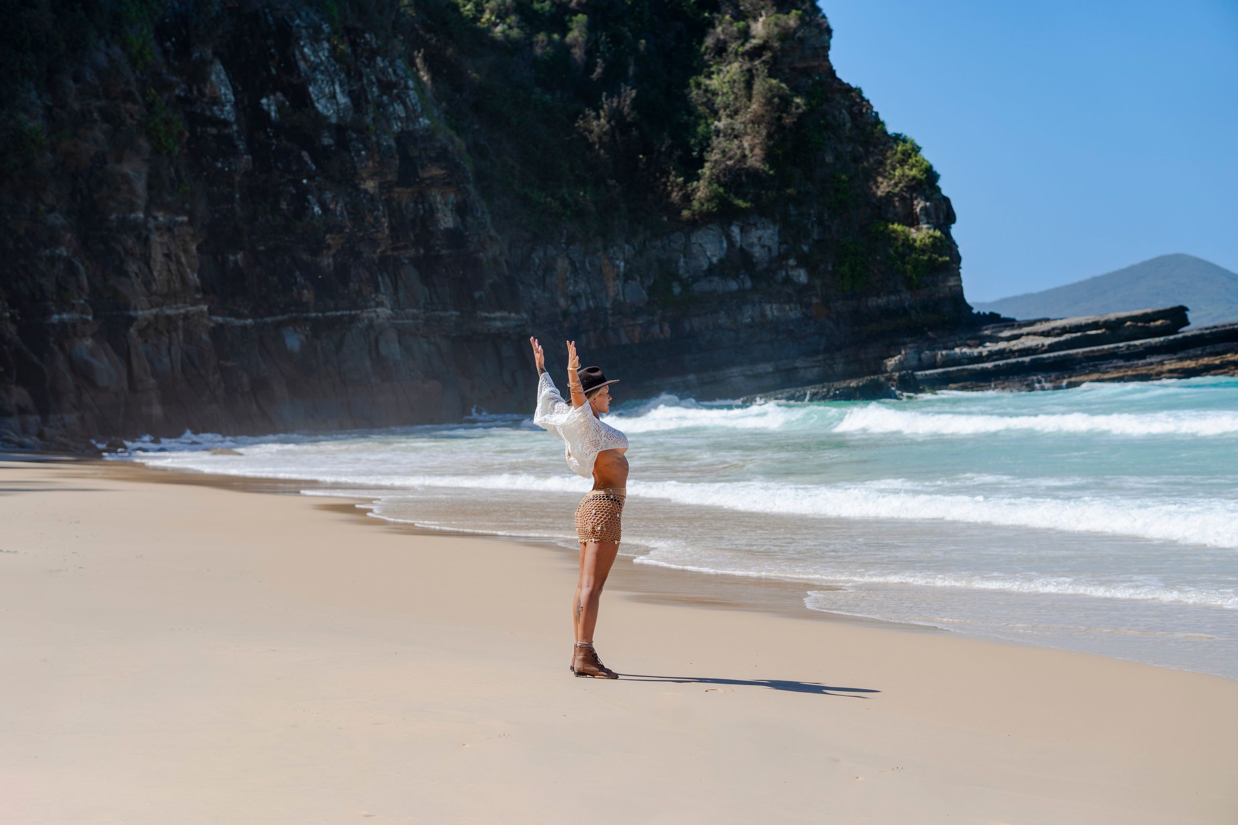 Woman in white top and shorts joyfully raises arms on a scenic sandy beach with rocky cliffs and ocean waves.