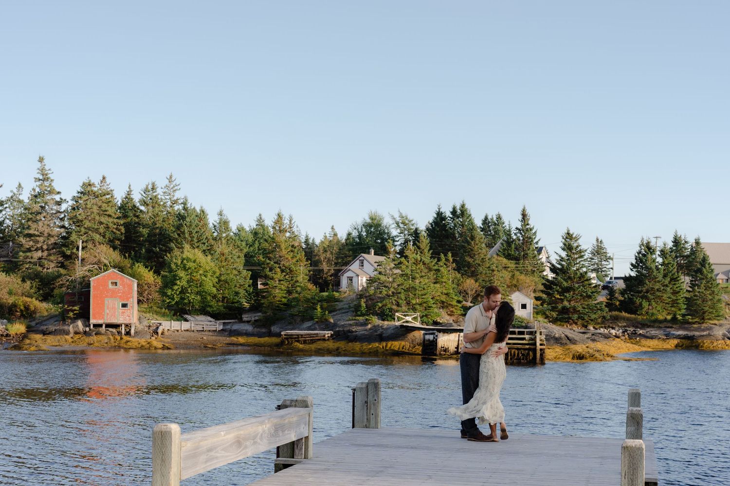 Couple embracing on a wooden dock overlooking calm water with evergreen trees and a red building in the background.