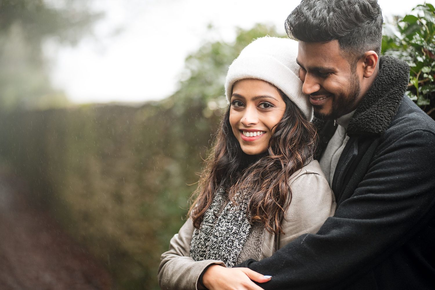 Happy couple embracing outdoors in winter, woman in white hat smiling, man in gray jacket, green hedge background.