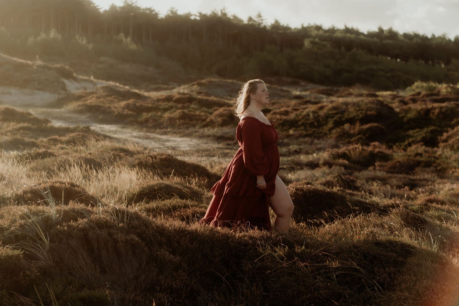 Woman in red dress standing on golden heathland, bathed in warm sunlight with forest backdrop.