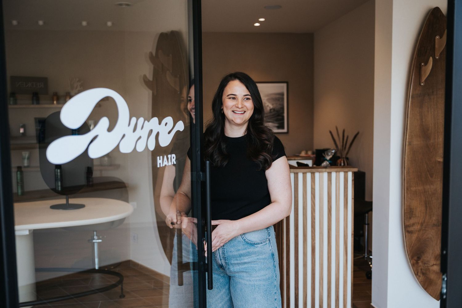 Woman smiling at entrance of Dune Hair salon, holding open glass door with logo, modern interior visible.