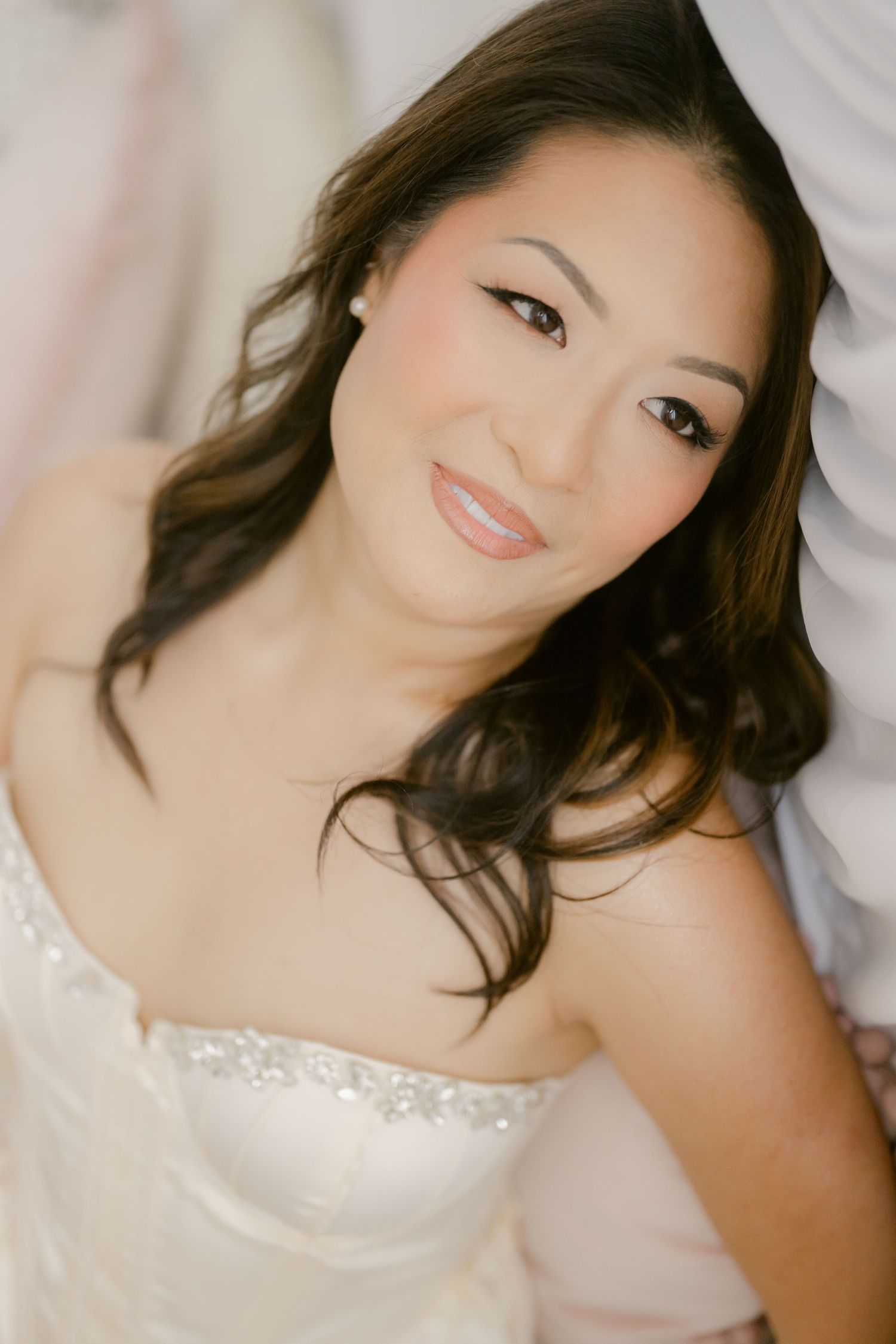 Beautiful Asian bride in strapless white wedding gown with beaded detail, wearing elegant makeup and pearl earrings.