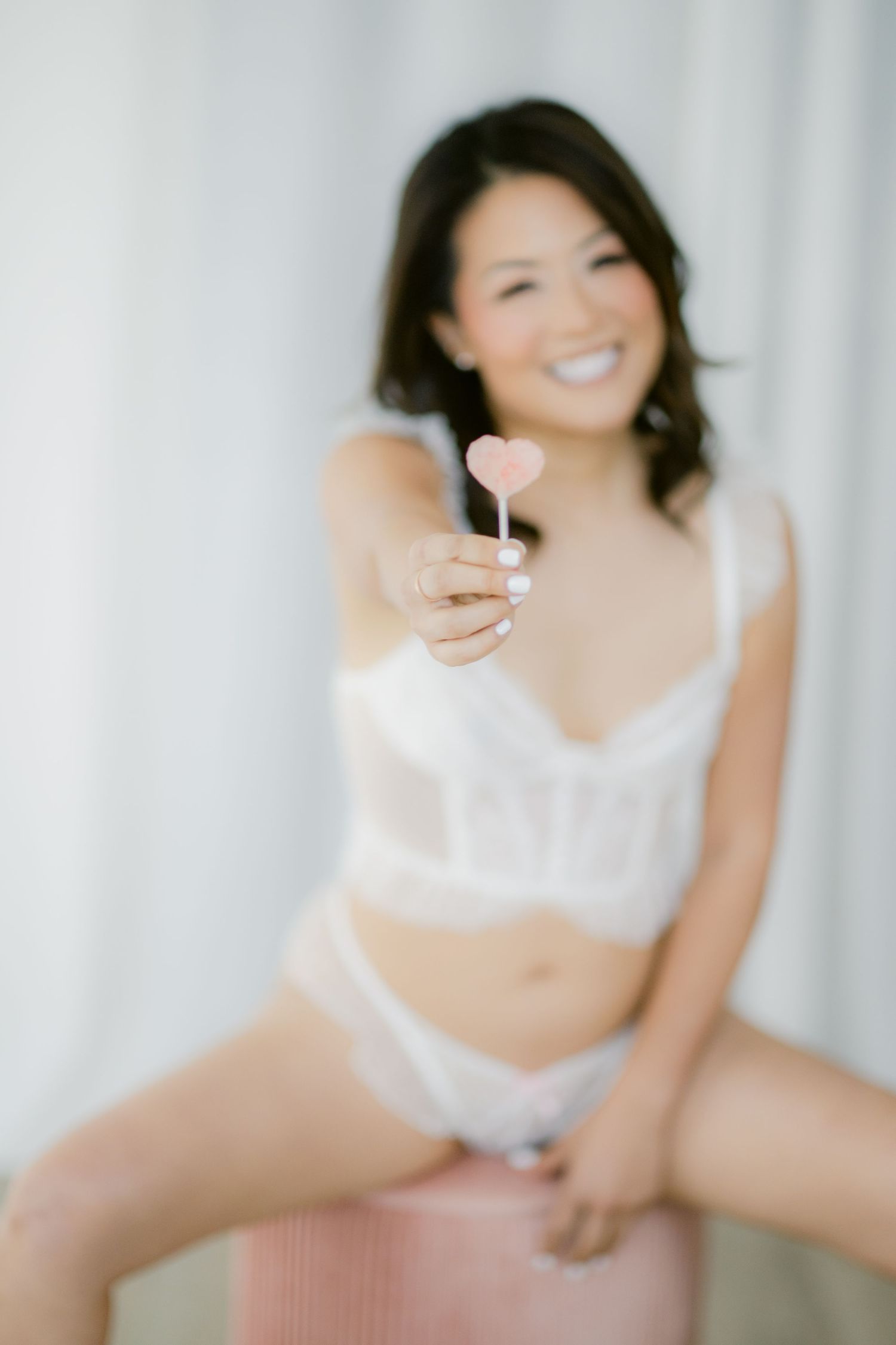 Smiling woman in white lingerie holding a pink heart-shaped lollipop toward the camera in a bright, airy setting.