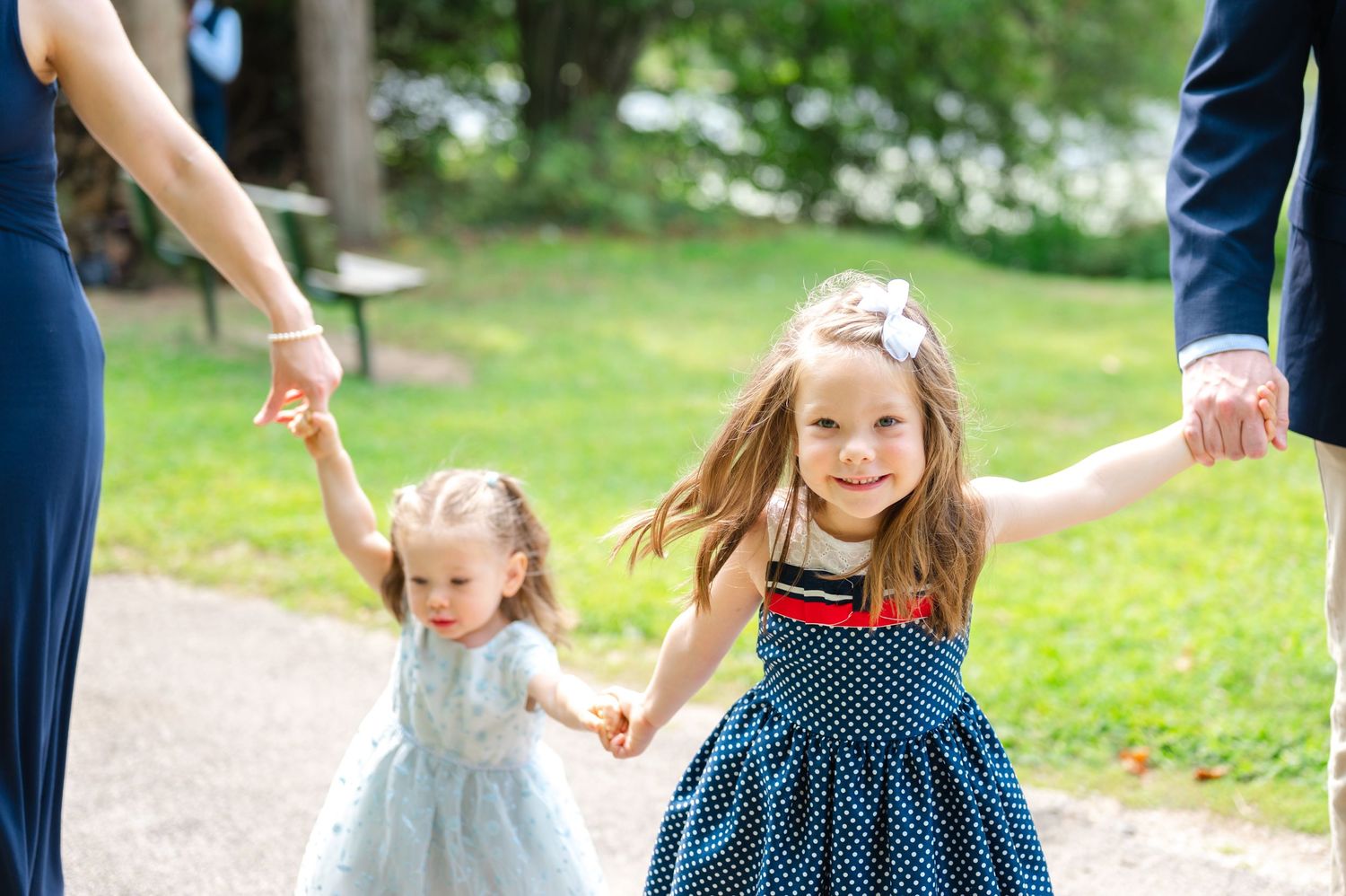 Two young girls in dresses hold hands with adults outdoors, smiling joyfully on a garden path.