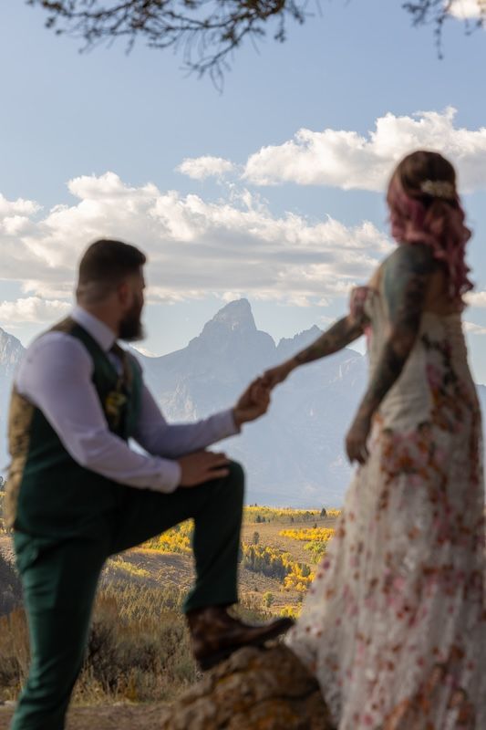 A groom kneels holding his tattooed bride's hand with pink hair in a floral dress, set against the Grand Teton mountains.