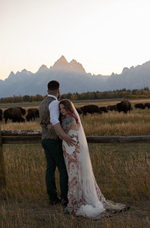 Couple in wedding attire embracing at sunset with Grand Teton mountains and bison grazing in background.