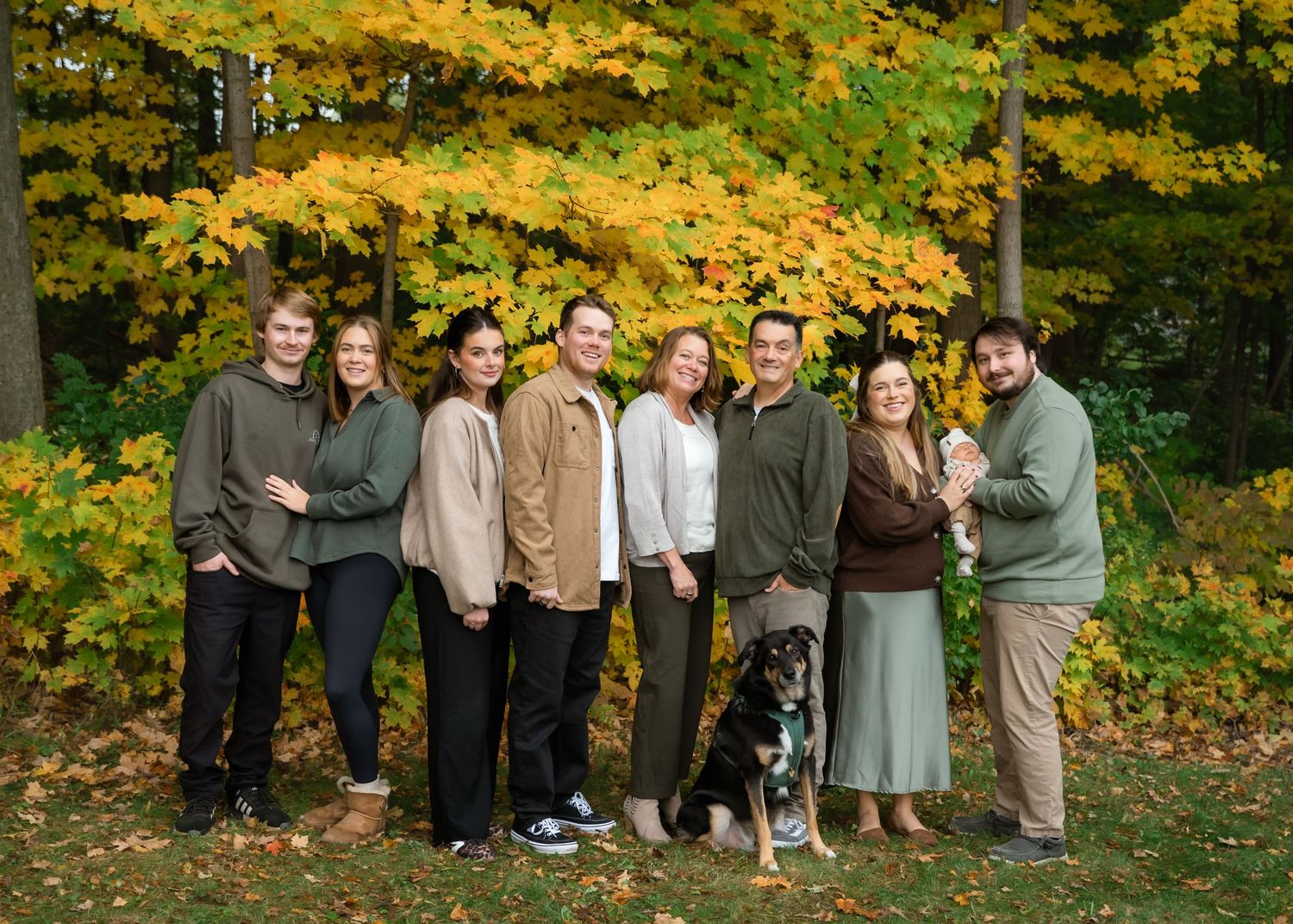 Group of eight people and a dog posing outdoors among vibrant autumn foliage in a lush green setting.