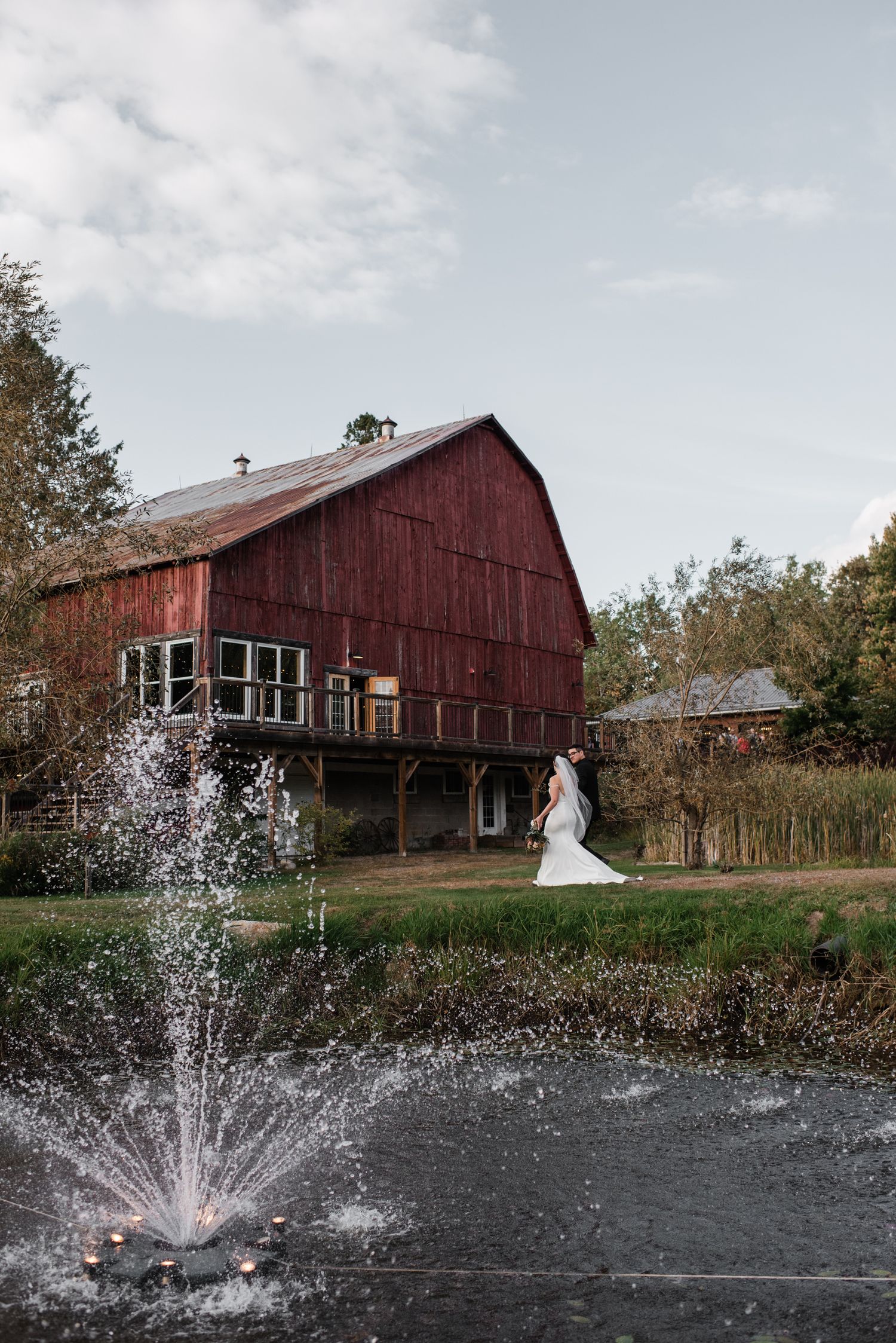 Bride in white gown stands a barn wedding venue Ontario exterior with a decorative pond fountain in the foreground.