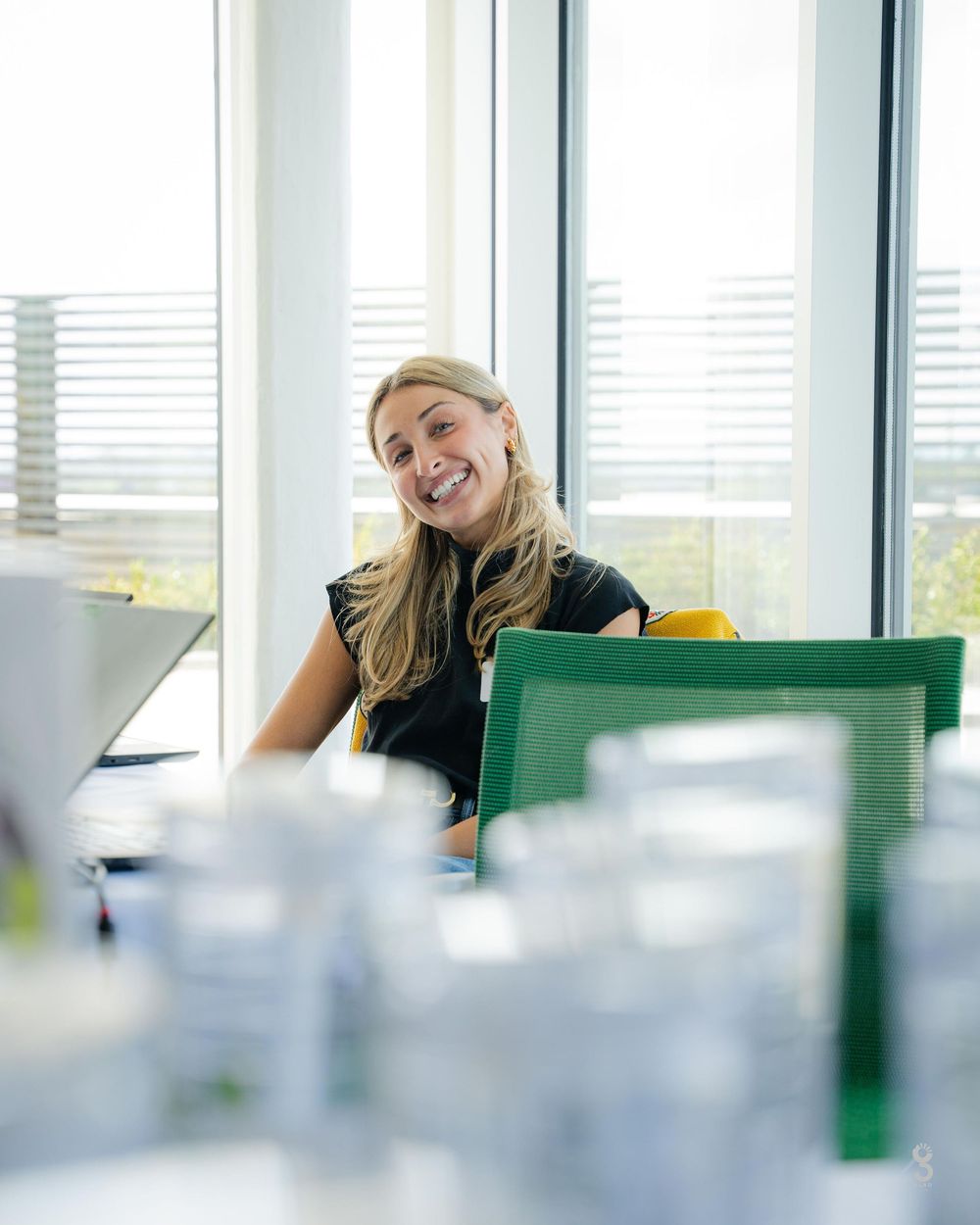 Professional sitting at bright modern office desk with windows and green chair visible in background.