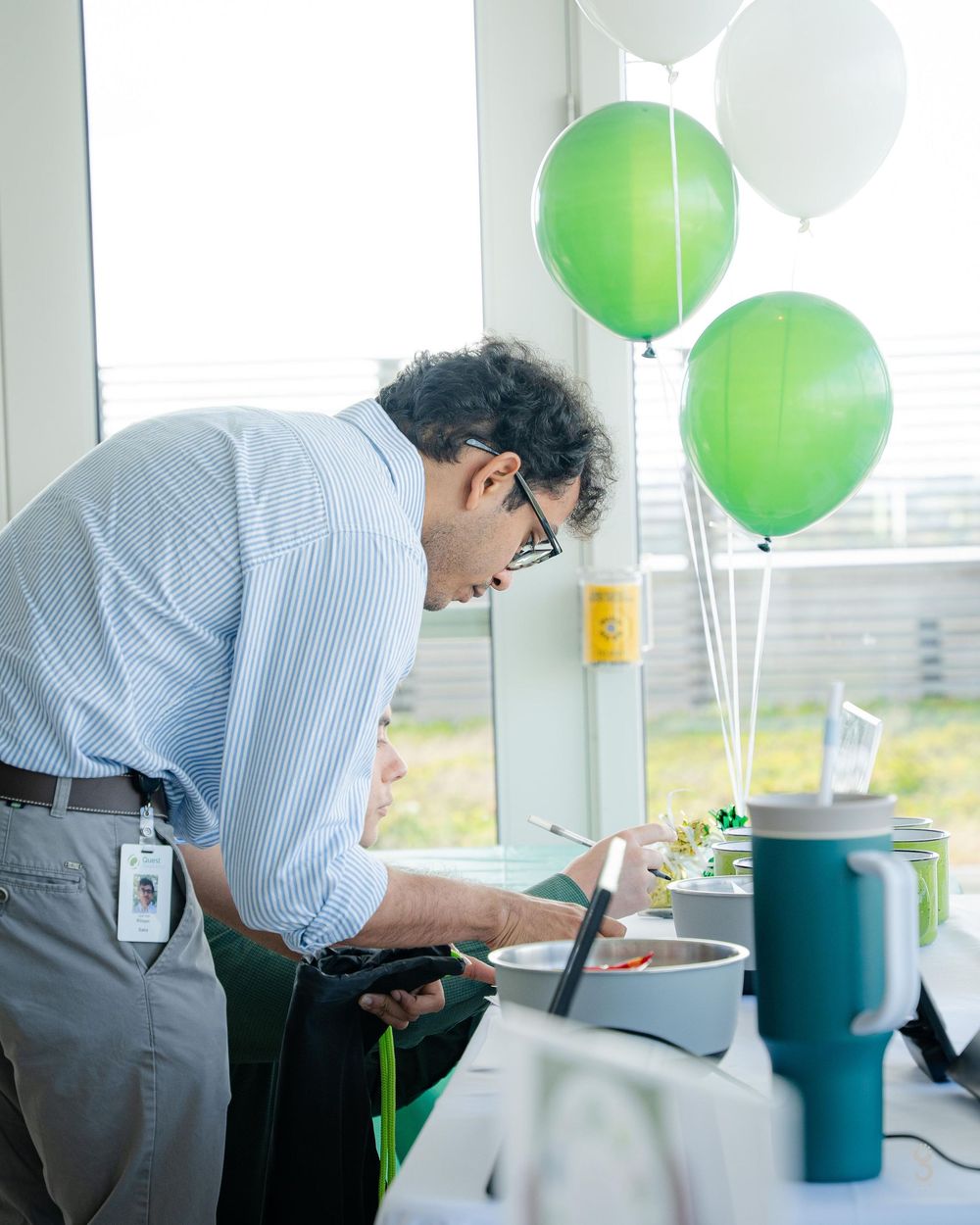 A person in a blue button-down shirt leans over a desk with green and white balloons in an office setting.