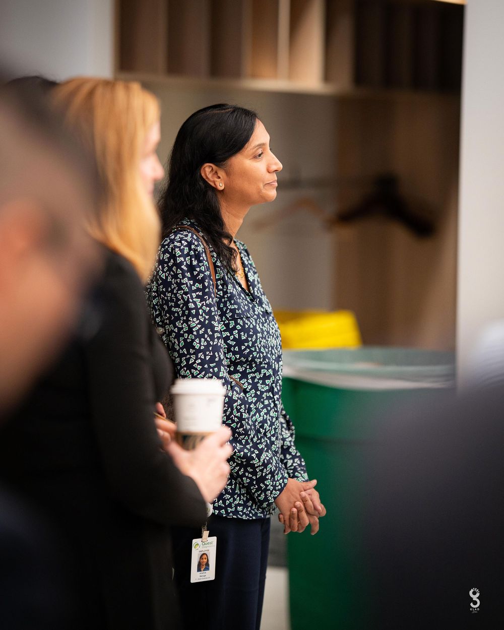Office workers with coffee cups gather in a modern workspace with green accents visible.