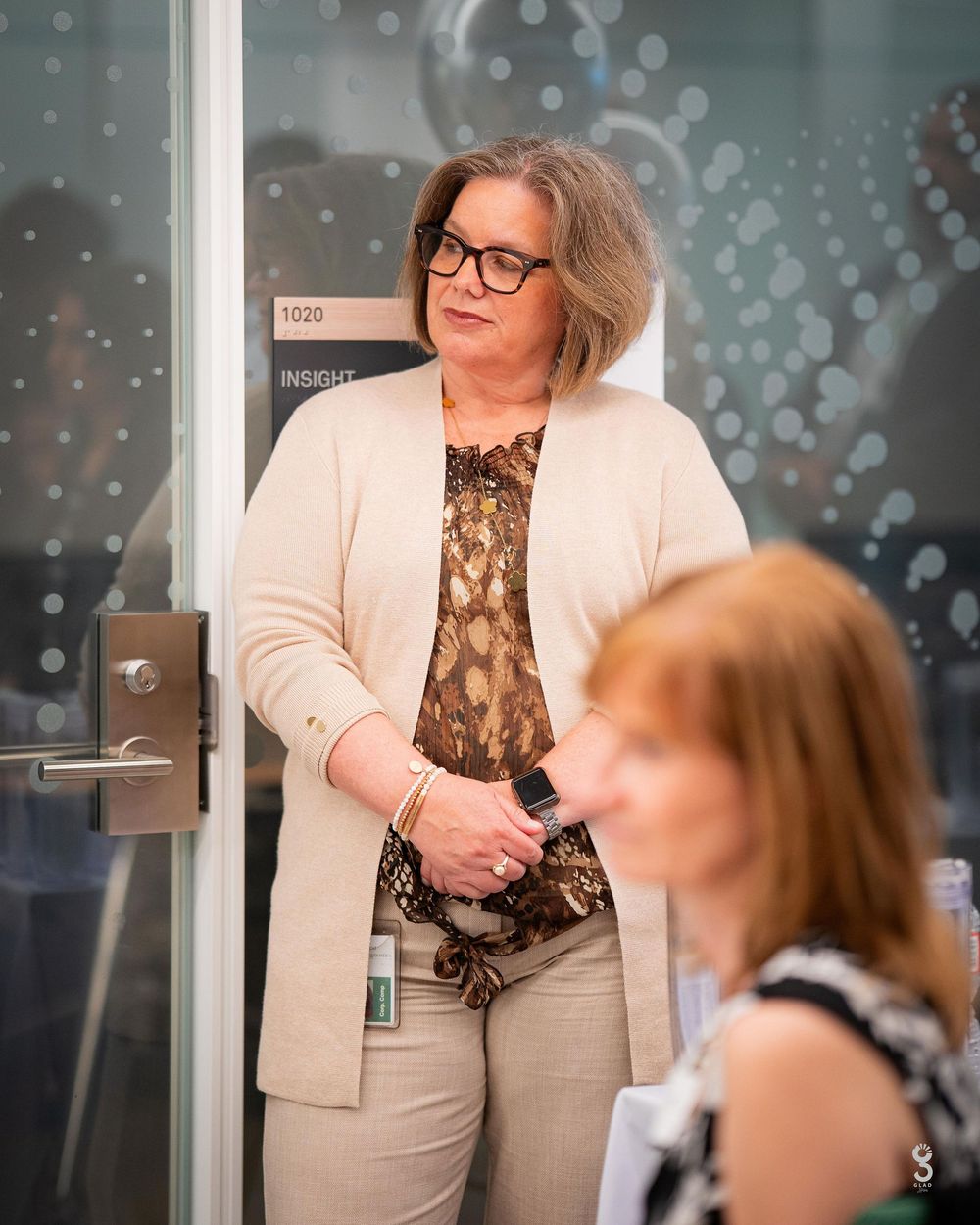 Professional wearing a beige suit and floral top interacts in an office setting with glass walls and bokeh lighting effects.