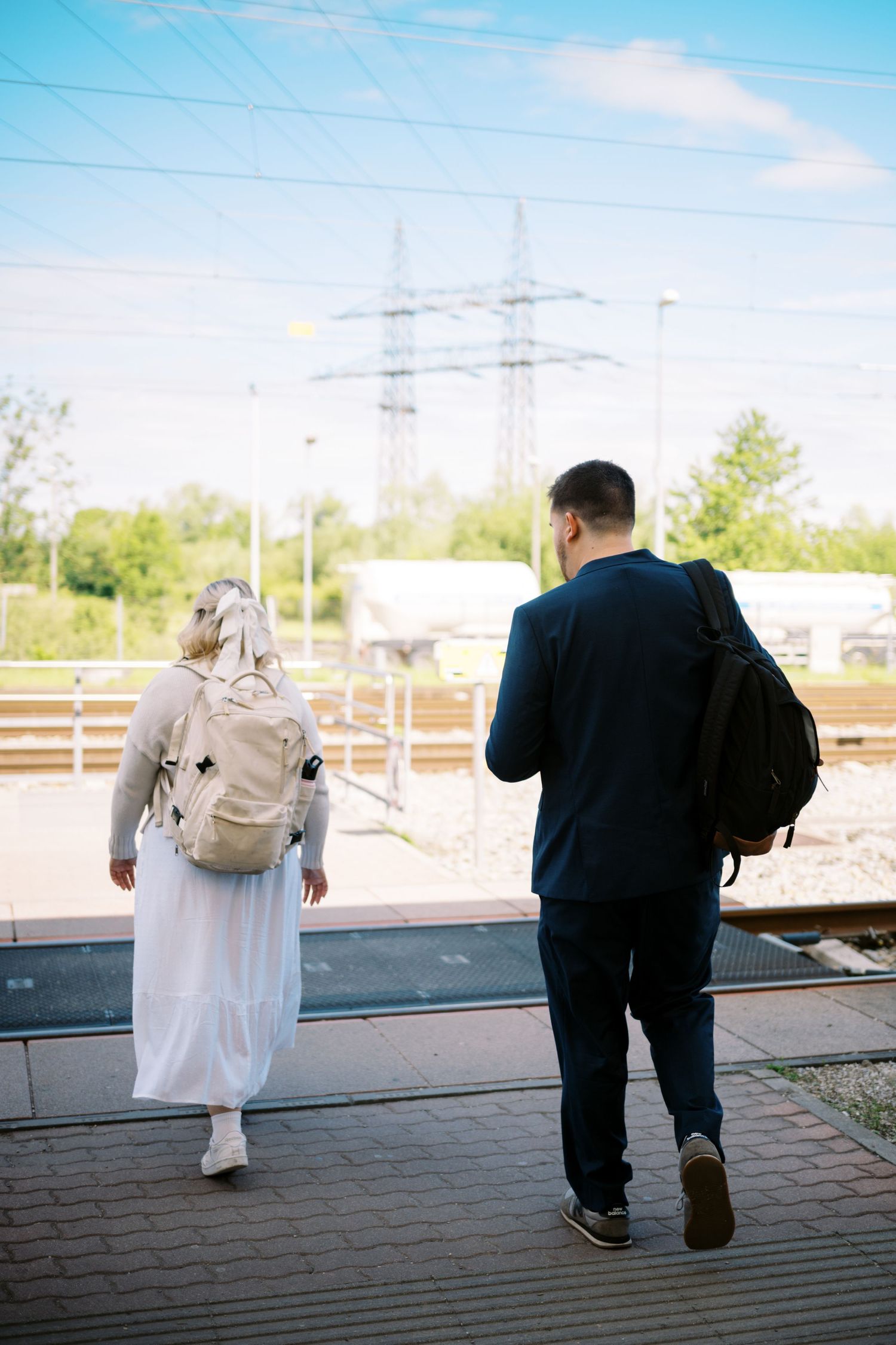 Two people carrying backpacks walking on a train platform with power lines visible in the background on a sunny day.