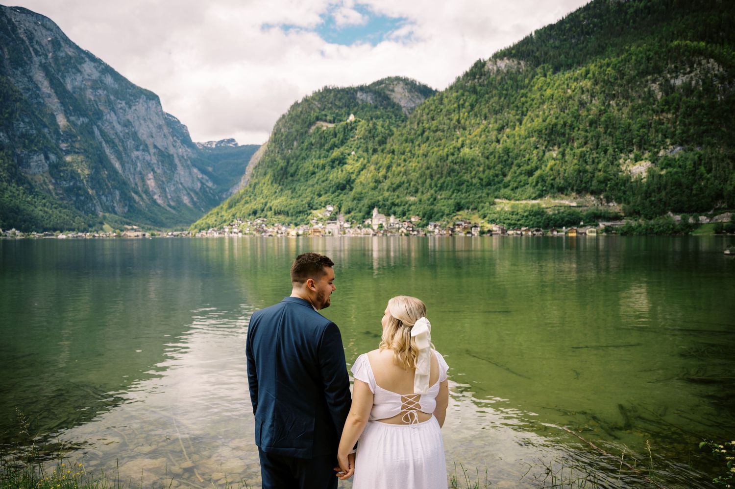 Two people stand by a serene mountain lake in Hallstatt, Austria with steep cliffs and village scenery in the background.