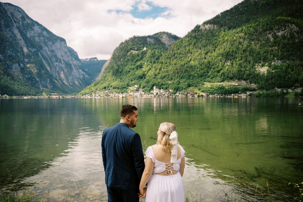 Two people stand by a serene mountain lake in Hallstatt, Austria with steep cliffs and village scenery in the background.
