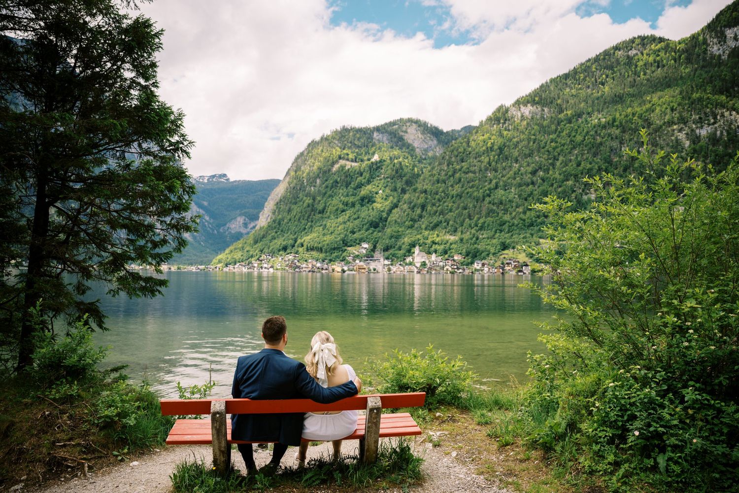 Two people sitting on bench overlooking serene lake surrounded by green mountains in Hallstatt.