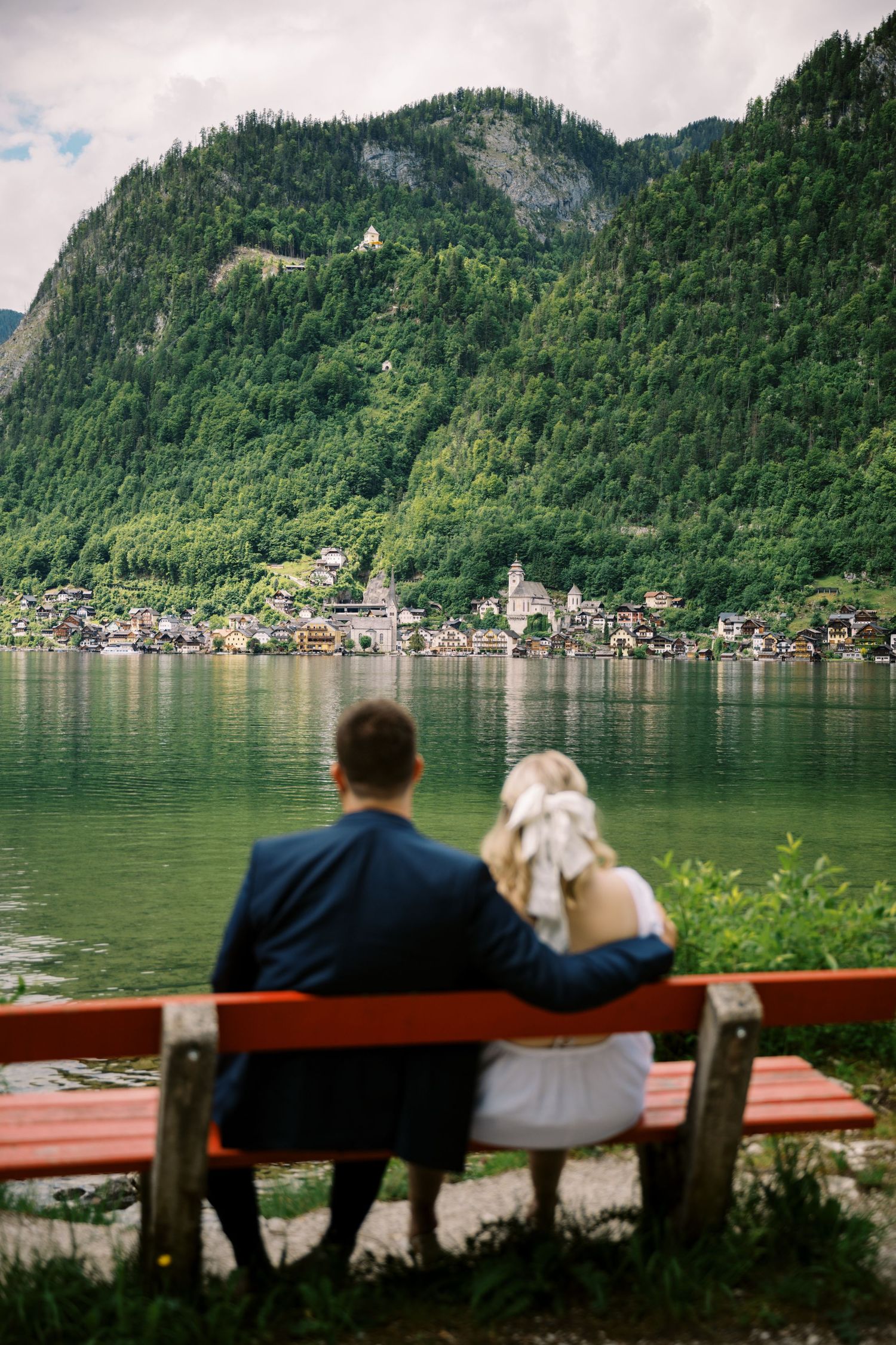 Two people sitting on a red bench overlooking a picturesque Alpine lake with mountains and village in background.