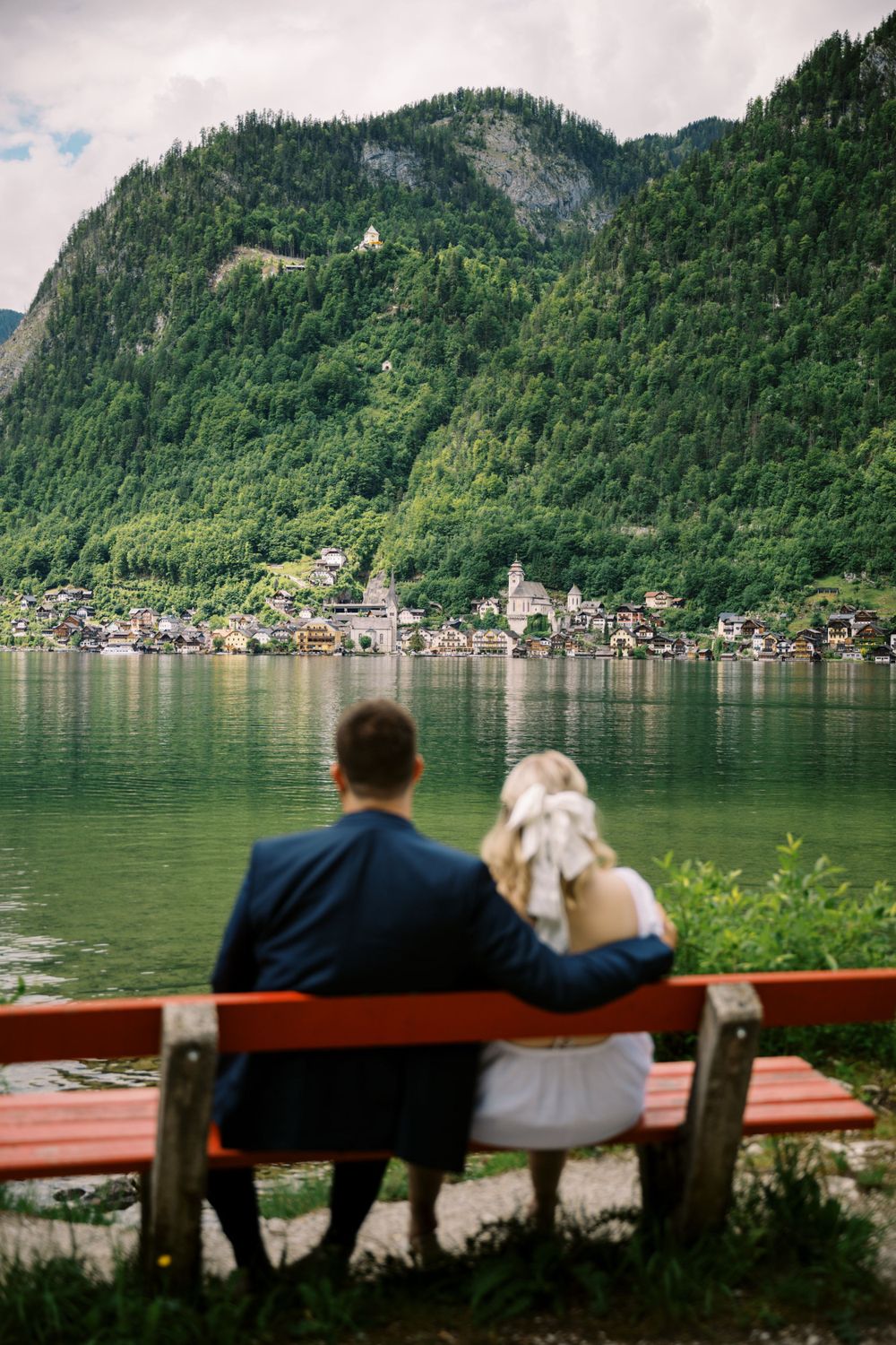 Two people sitting on a red bench overlooking a picturesque Alpine lake with mountains and village in background.
