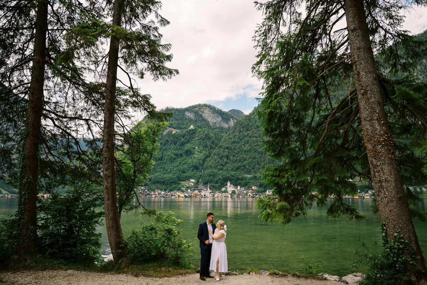 Lake view through pine trees in Hallstatt Austria with two people standing at waters edge in romantic embrace.