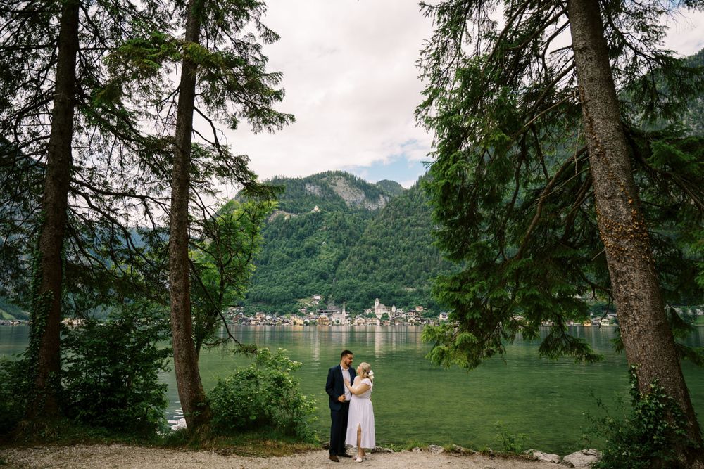 Lake view through pine trees in Hallstatt Austria with two people standing at waters edge in romantic embrace.