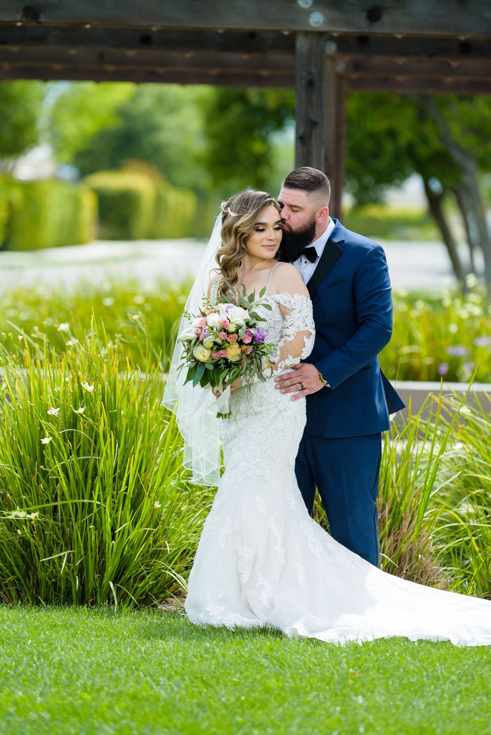 Wedding couple embraces in lush green garden setting with bridal bouquet, bride in lace gown and groom in navy suit.