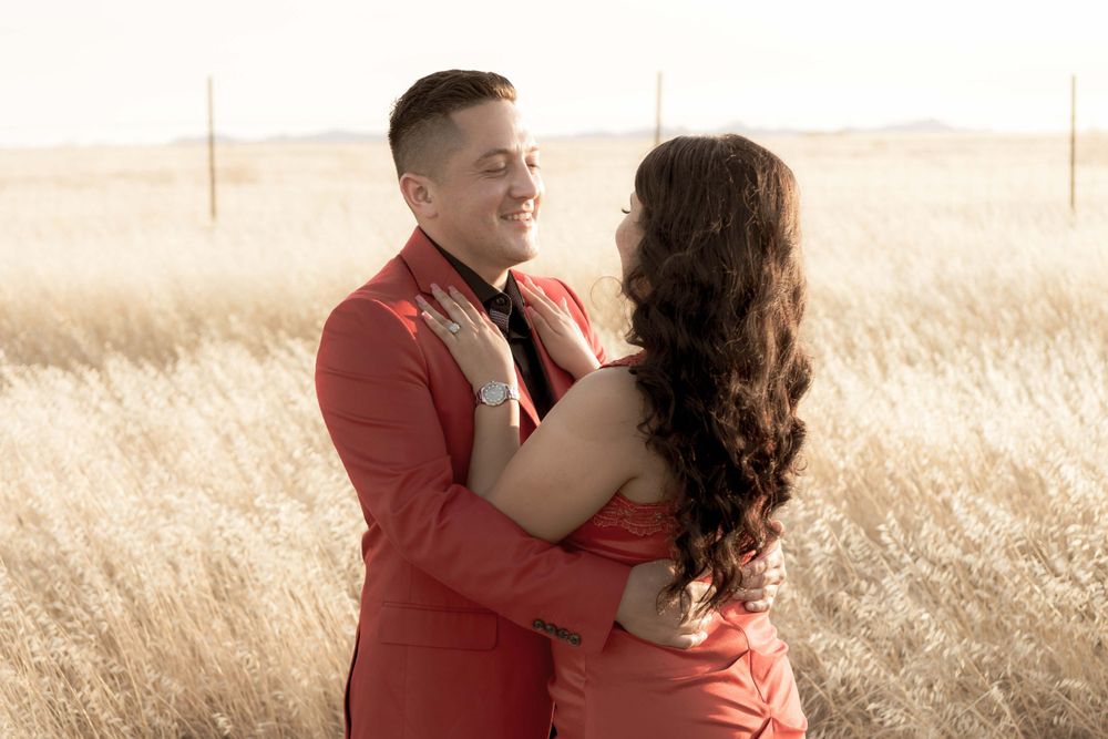 A couple embraces in warm sunlight while standing in a golden wheat field wearing matching coral outfits.