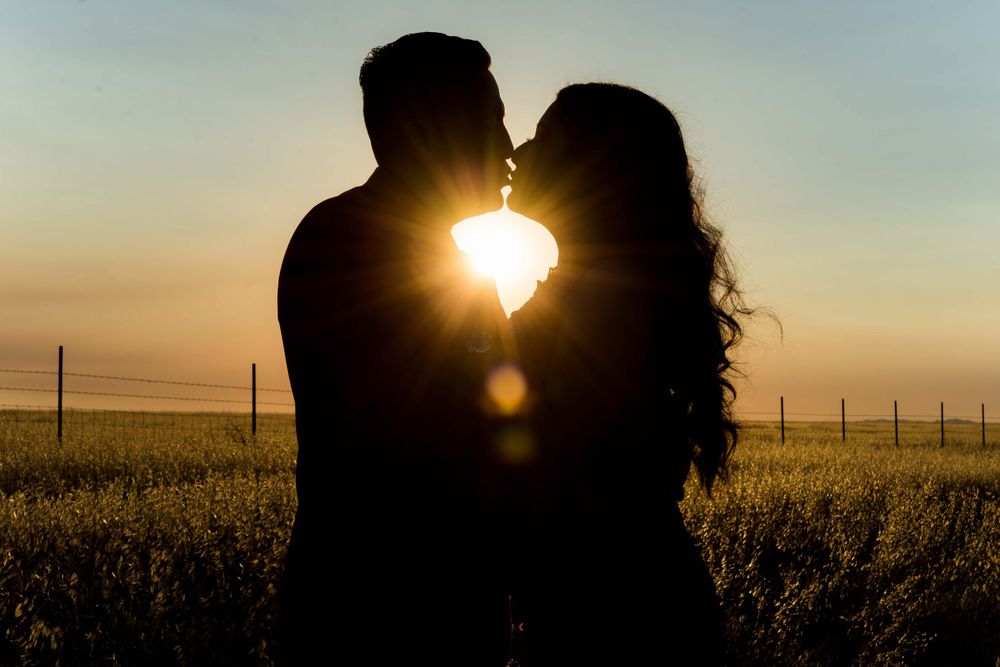 Silhouetted couple sharing romantic kiss at sunset with sunburst between them in rural field.