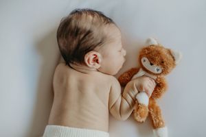 A sleeping baby lies peacefully next to a soft brown teddy bear on white bedding.