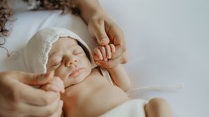 A gentle hand touches a sleeping newborn baby wearing a delicate white lace bonnet.