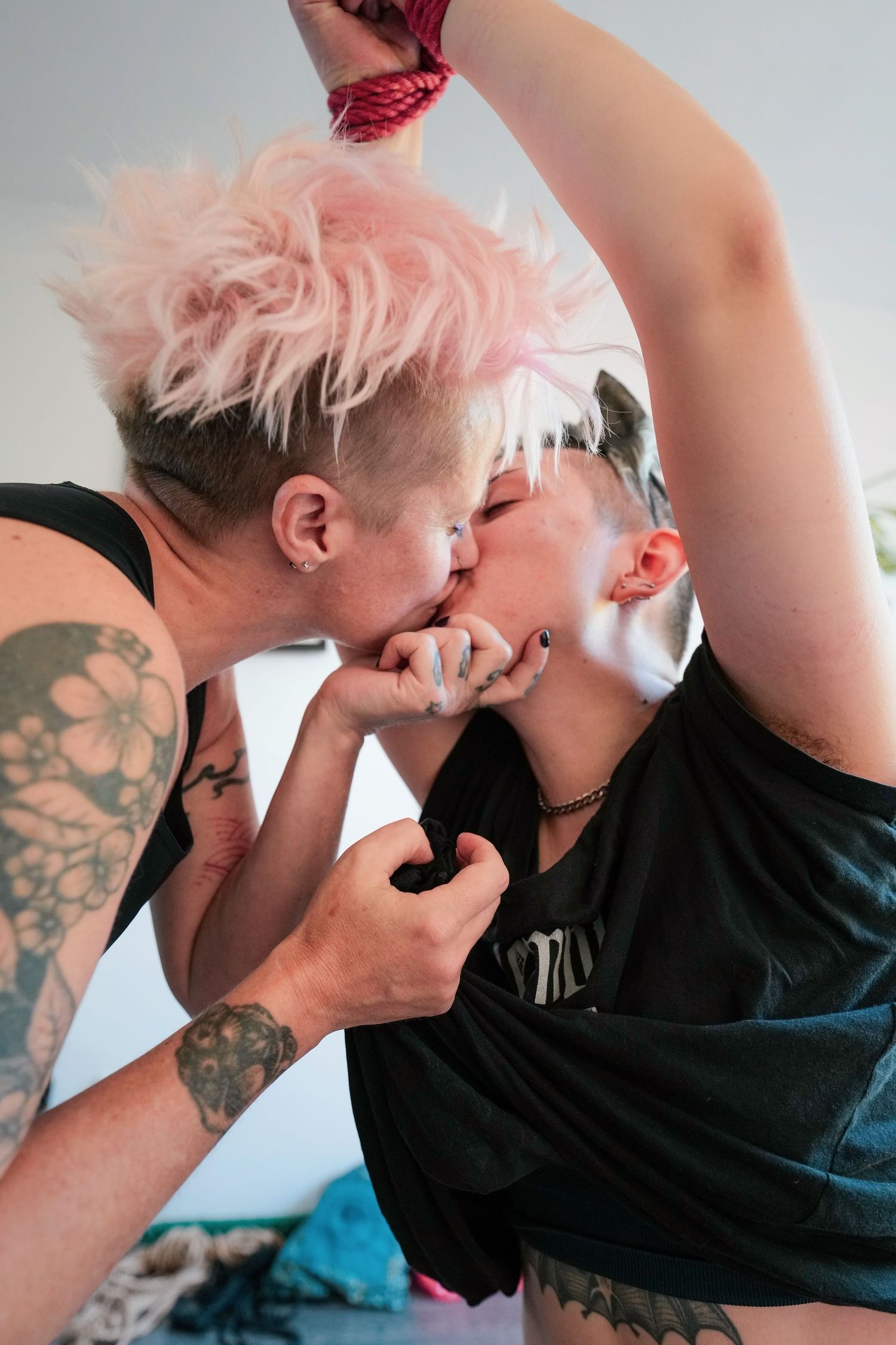 Two people sharing an intimate moment against a light background with visible tattoos and colorful hair.