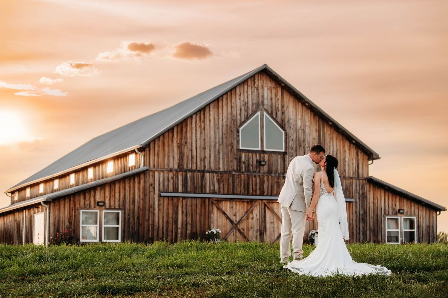 A wedding couple stands silhouetted against a rustic barn at sunset with warm orange sky.