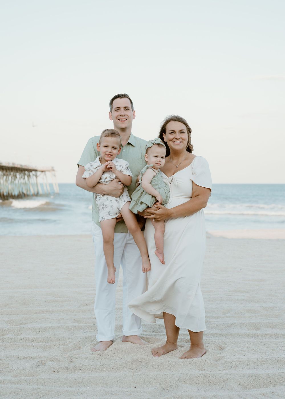 Family of four poses on beach at sunset, parents holding two young children in coordinating sage green and white outfits.