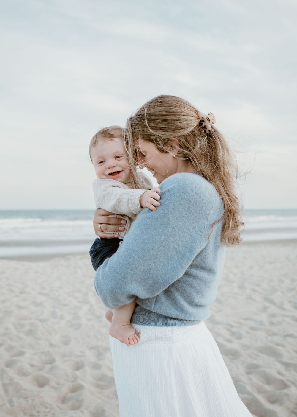 Mother in blue sweater and white skirt holds laughing baby boy on sandy beach at dusk.