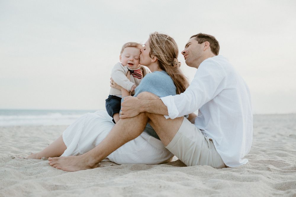Young family sits together on beach sand, baby boy laughing joyfully as parents share a kiss.