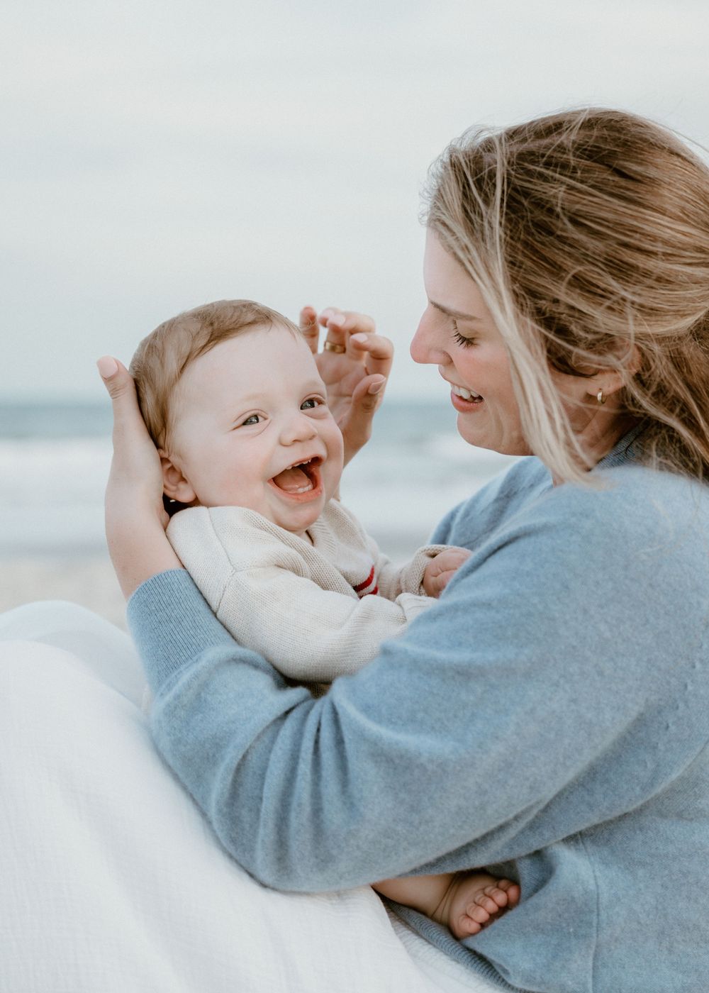 Smiling baby boy laughs while mother in blue sweater holds him on overcast beach.