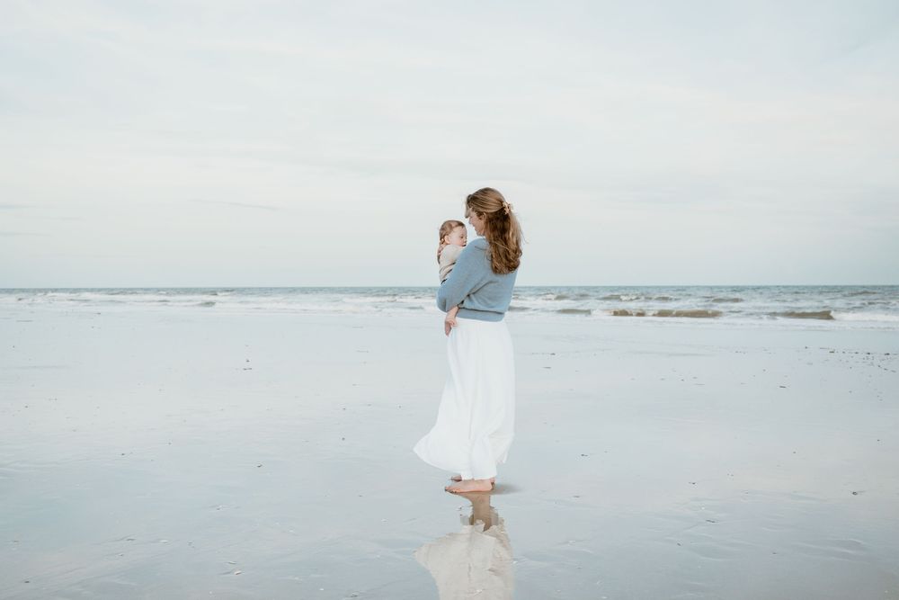 Mother in blue sweater and white skirt holds baby while standing at ocean's edge on cloudy beach.