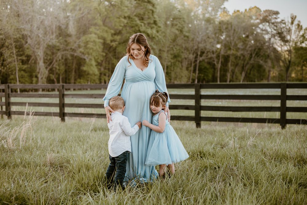 Family wearing complementary blue outfits poses together in grassy field with wooden fence backdrop at sunset.