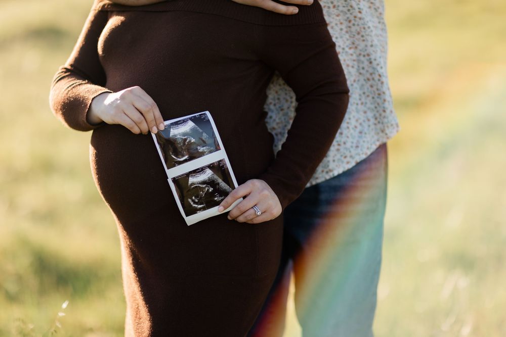 A pregnant person in a black dress holds ultrasound photos while standing in sunlight with a rainbow lens flare effect.