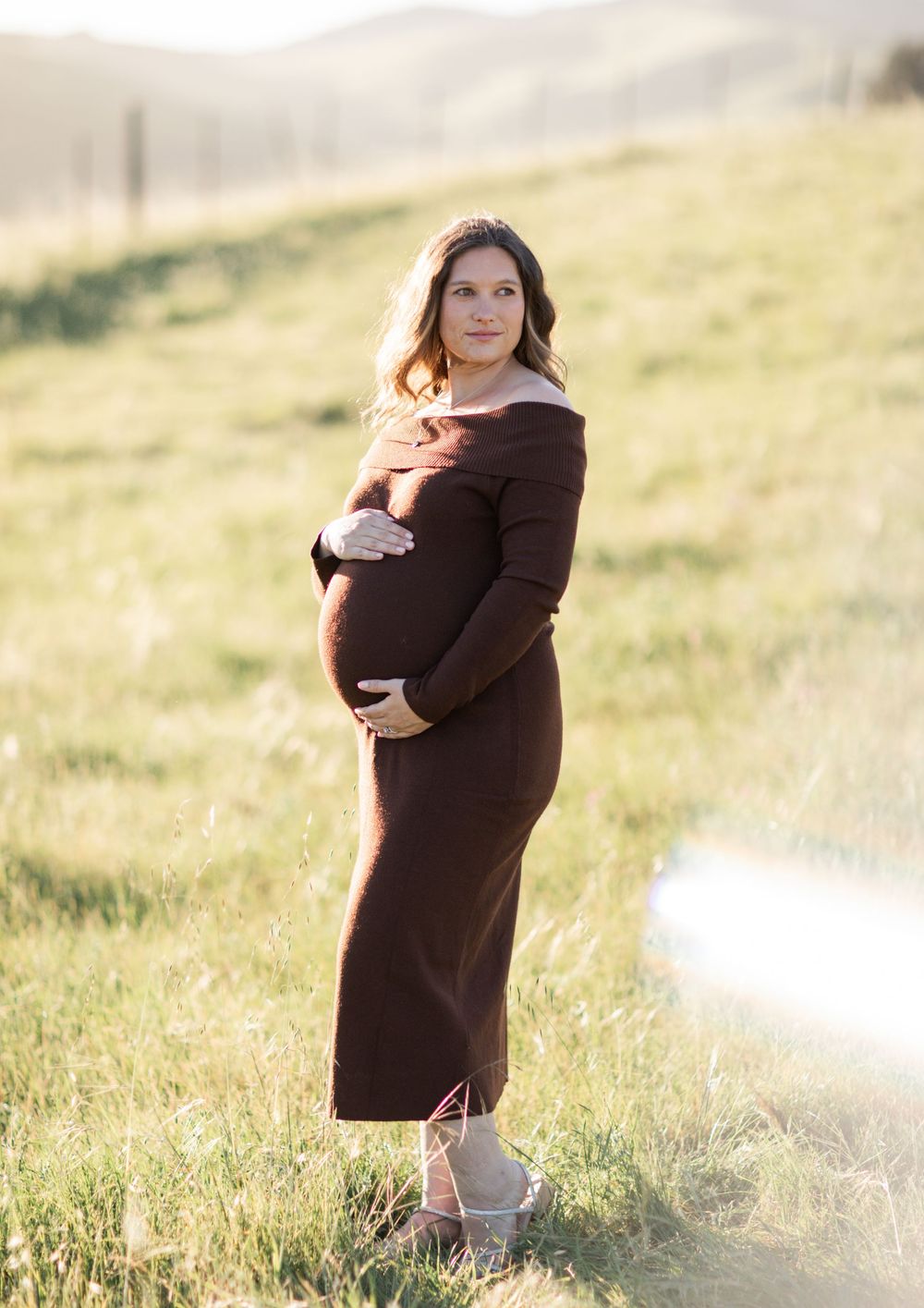 Maternity photo session in a sunlit meadow with soft golden light filtering through tall grass.