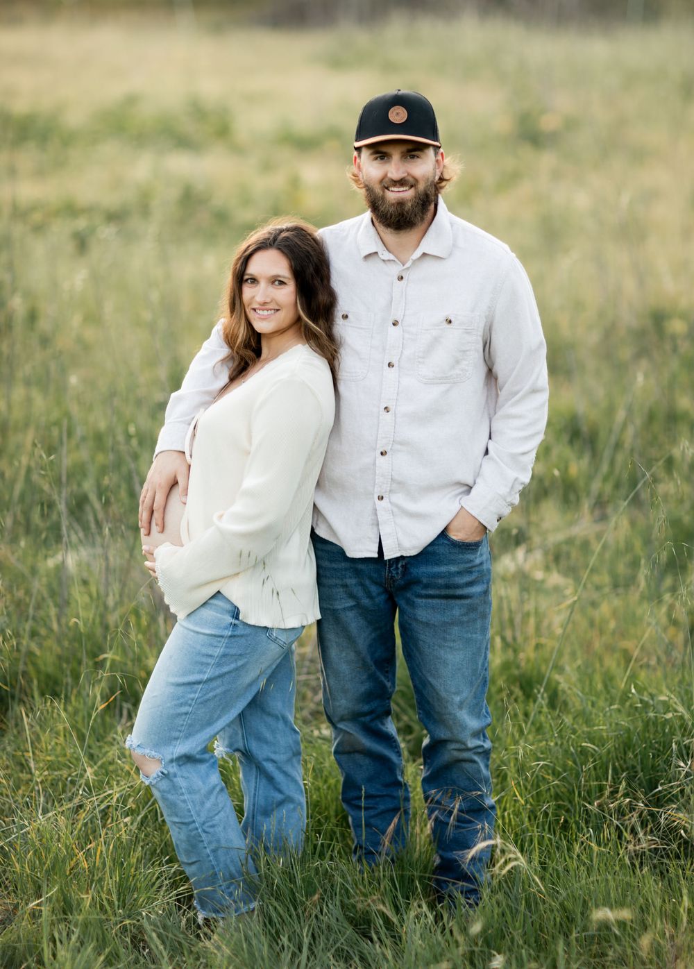 A couple in casual attire embraces while standing in a grassy meadow on a sunny day.