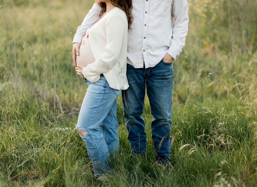 A couple in white shirts and blue jeans stands together in tall green grass during an outdoor photoshoot.