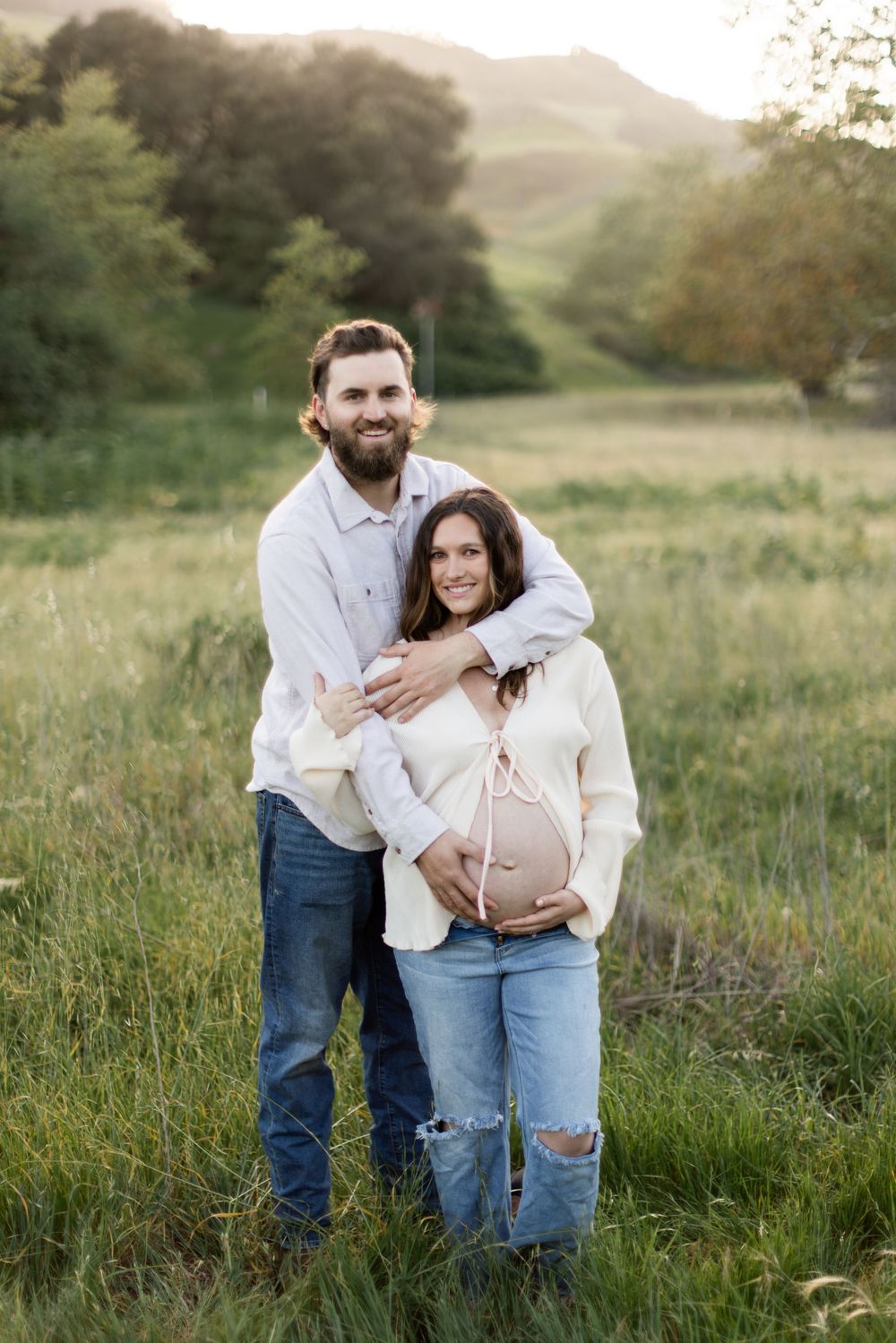 A couple embraces in a sunlit meadow with rolling hills and green grass in the background during golden hour.