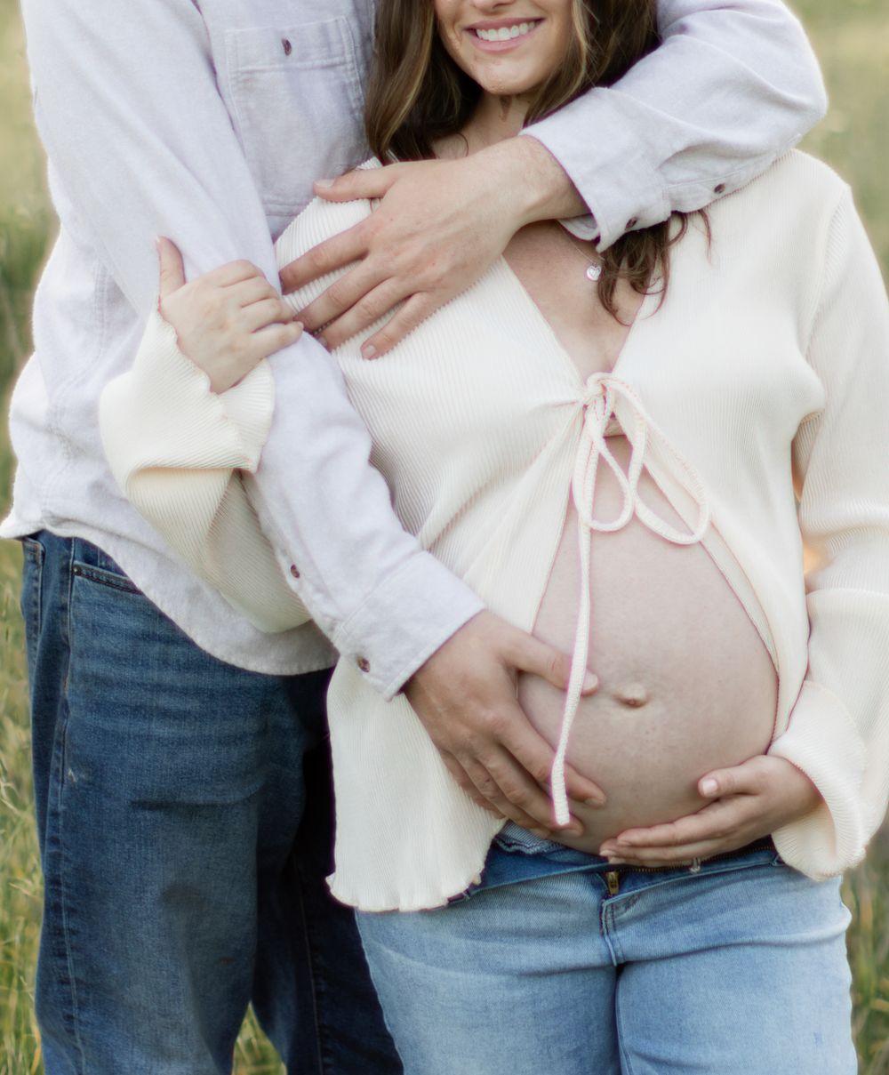 Couple embraces during maternity photo shoot with hands resting on pregnant belly wearing jeans and white sweater outdoors.