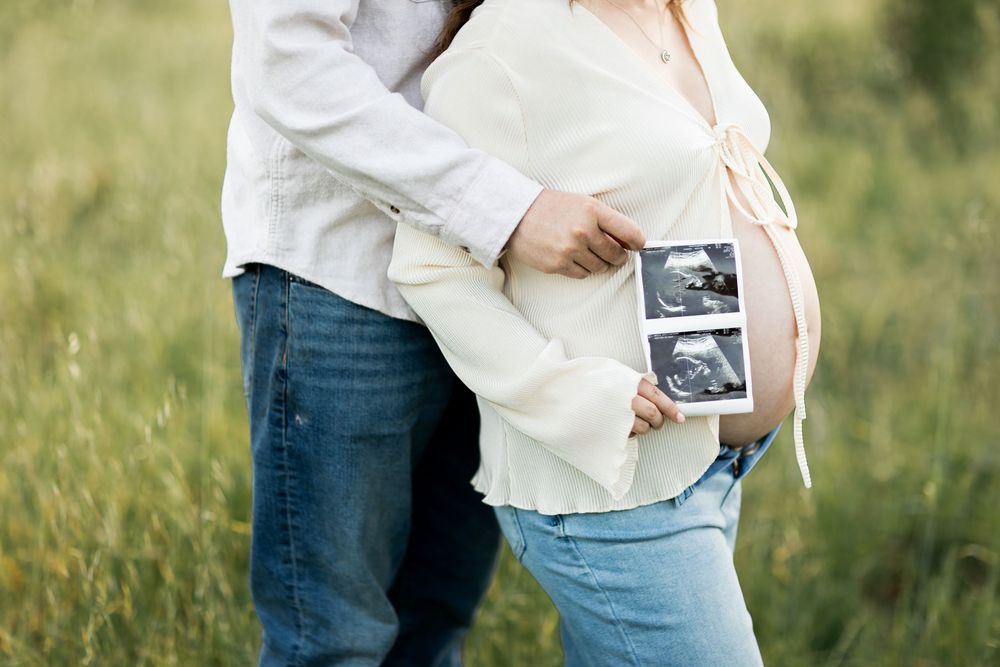 A couple in casual attire holds a sonogram photo while standing together in a sunlit grassy field.