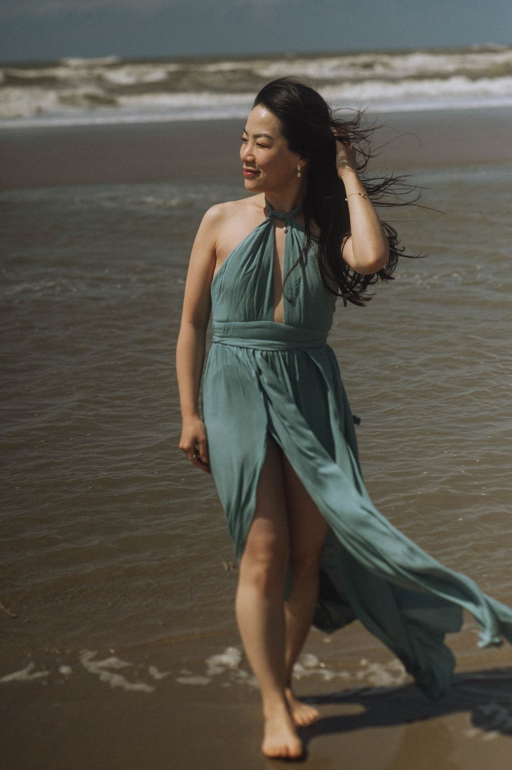 Woman in teal dress dances playfully along the beach shoreline.