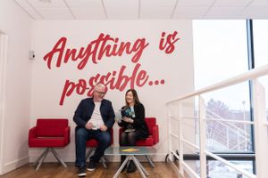 Two people sit on a red couch beneath red wall text reading 'Anything is possible' in modern office space with windows.
