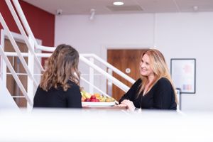 Two women having a professional conversation at a modern white desk with a bowl of fruit between them.
