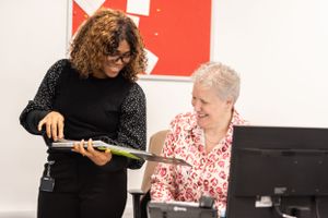 Two people discussing while looking at a tablet device in an office setting with red wall background.