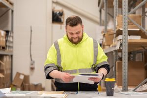 Warehouse worker in high visibility safety vest reviews paperwork while standing between storage racks.