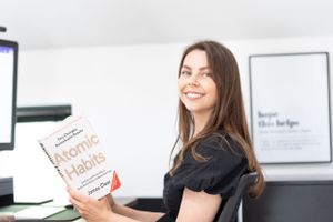 Someone smiling while holding up a copy of Atomic Habits book in an office setting with white walls and computer screens.